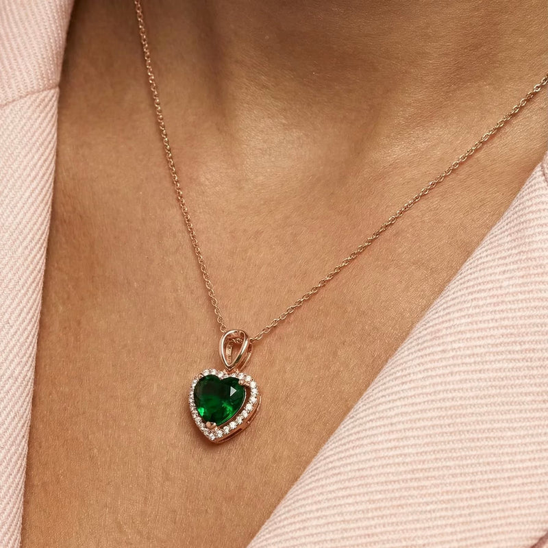 Gold-plated heart pendant necklace with an emerald-green center stone and a pavé halo, worn at the neckline.